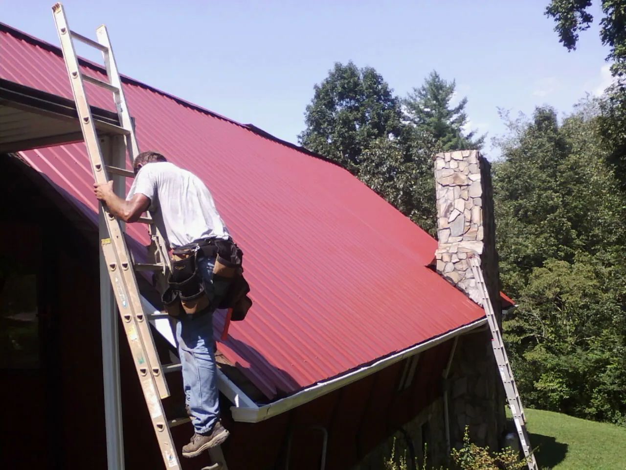Roofer on a ladder beside a red metal roof and stone chimney — Soffit & Fascia Repair by Roofing Brothers in Friedens